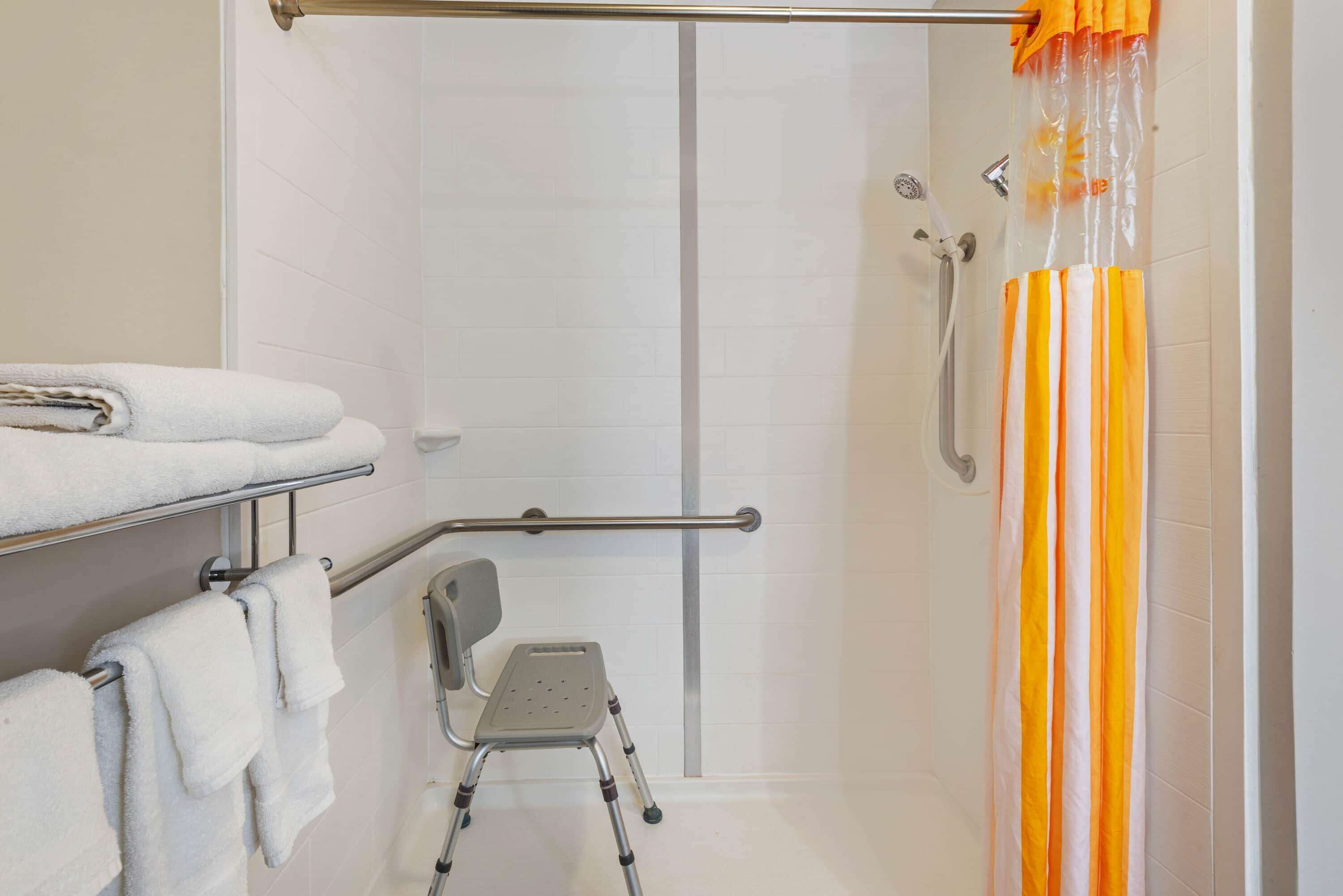 A bathroom with a shower, a grab bar, a wheelchair-accessible bench, and a towel rack with folded towels.