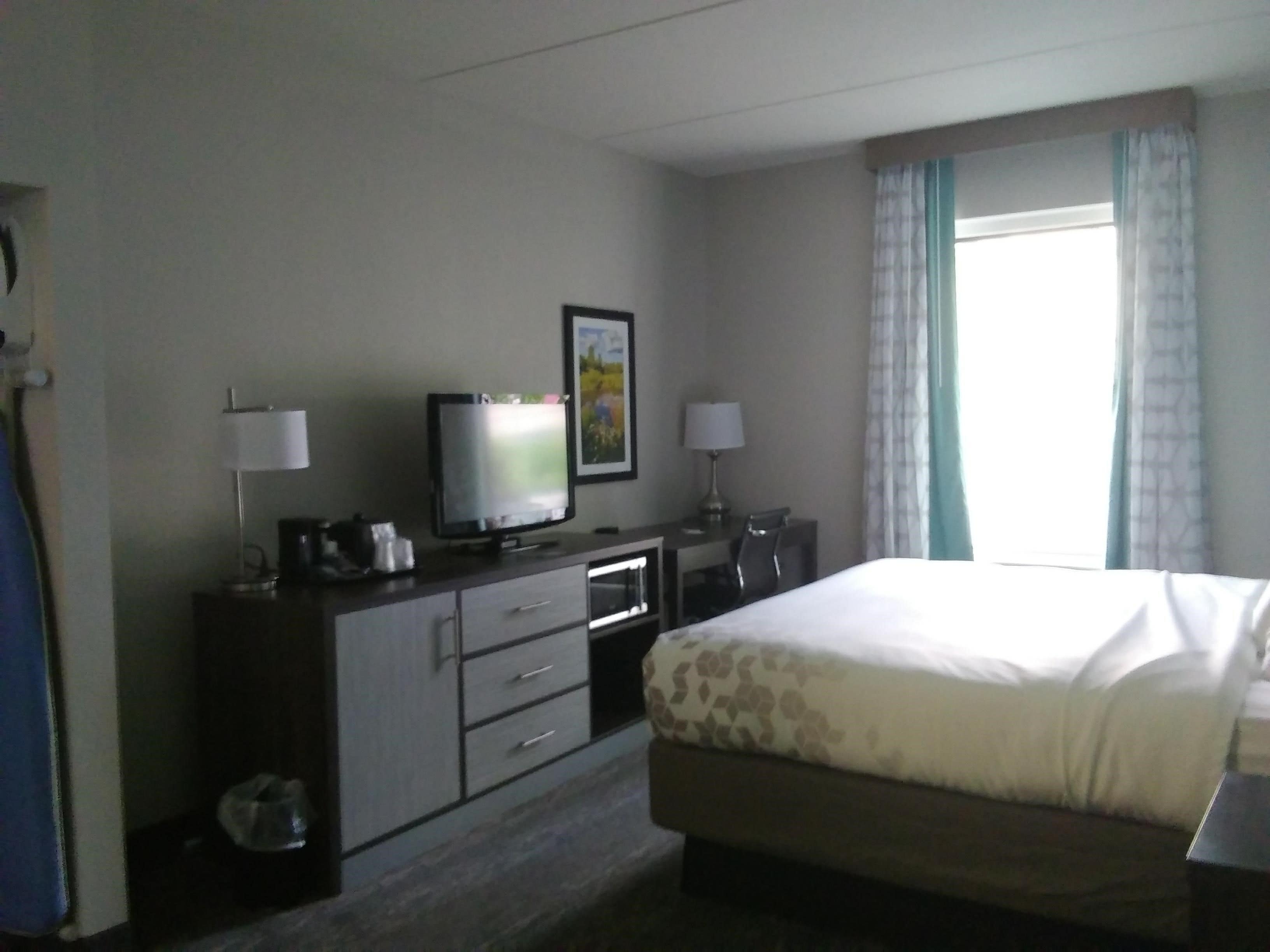 A hotel room with a bed, a television, a desk with a lamp, and a window with curtains.