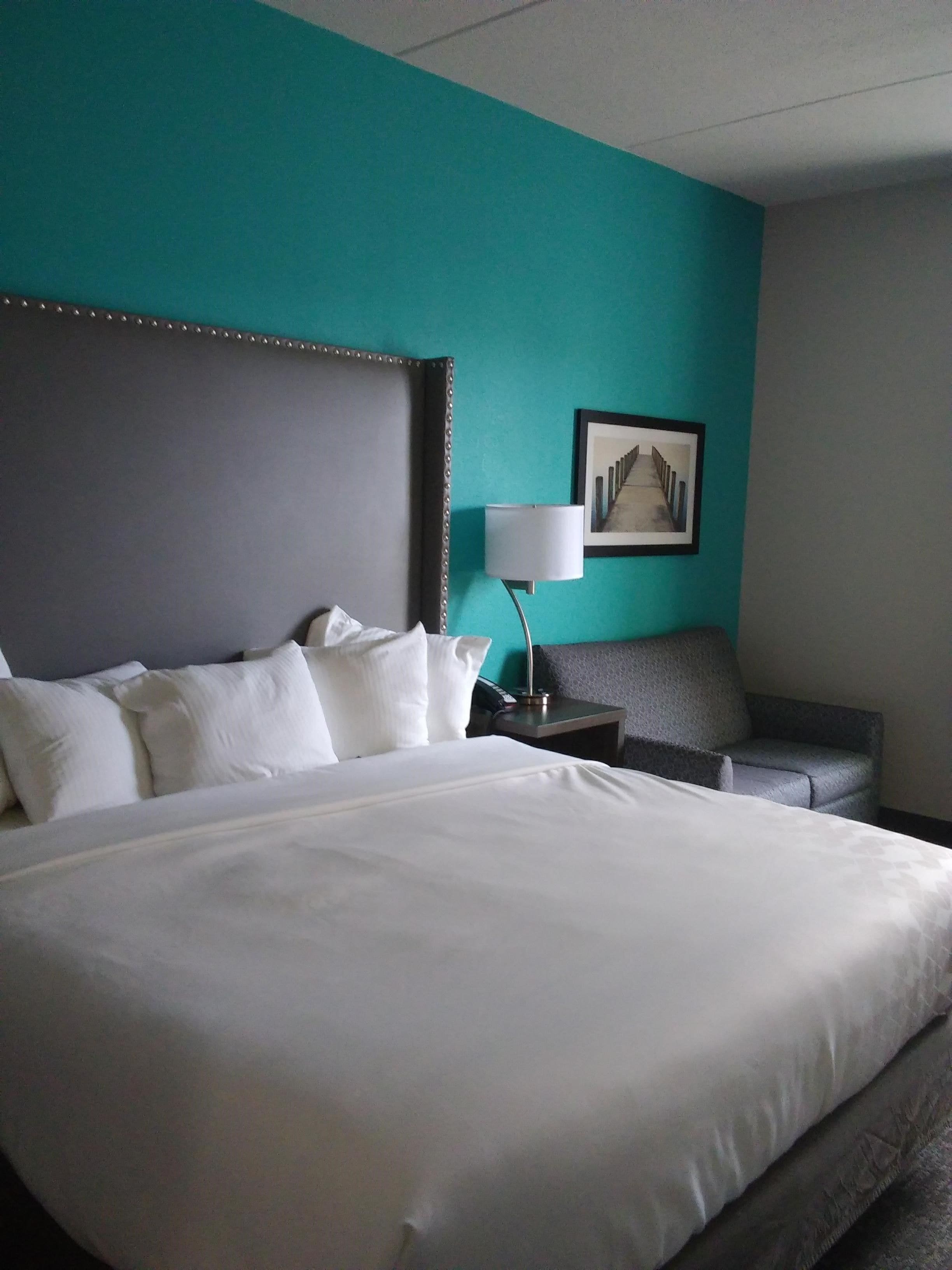 A hotel room with a large bed, a bedside table with a lamp, a grey armchair, and a framed picture on the wall.