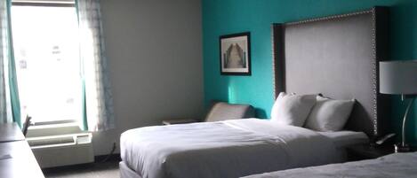 Habitación de hotel con dos camas, un escritorio, una silla, una ventana con cortinas, un cuadro en la pared y una mesita de noche con lámpara.