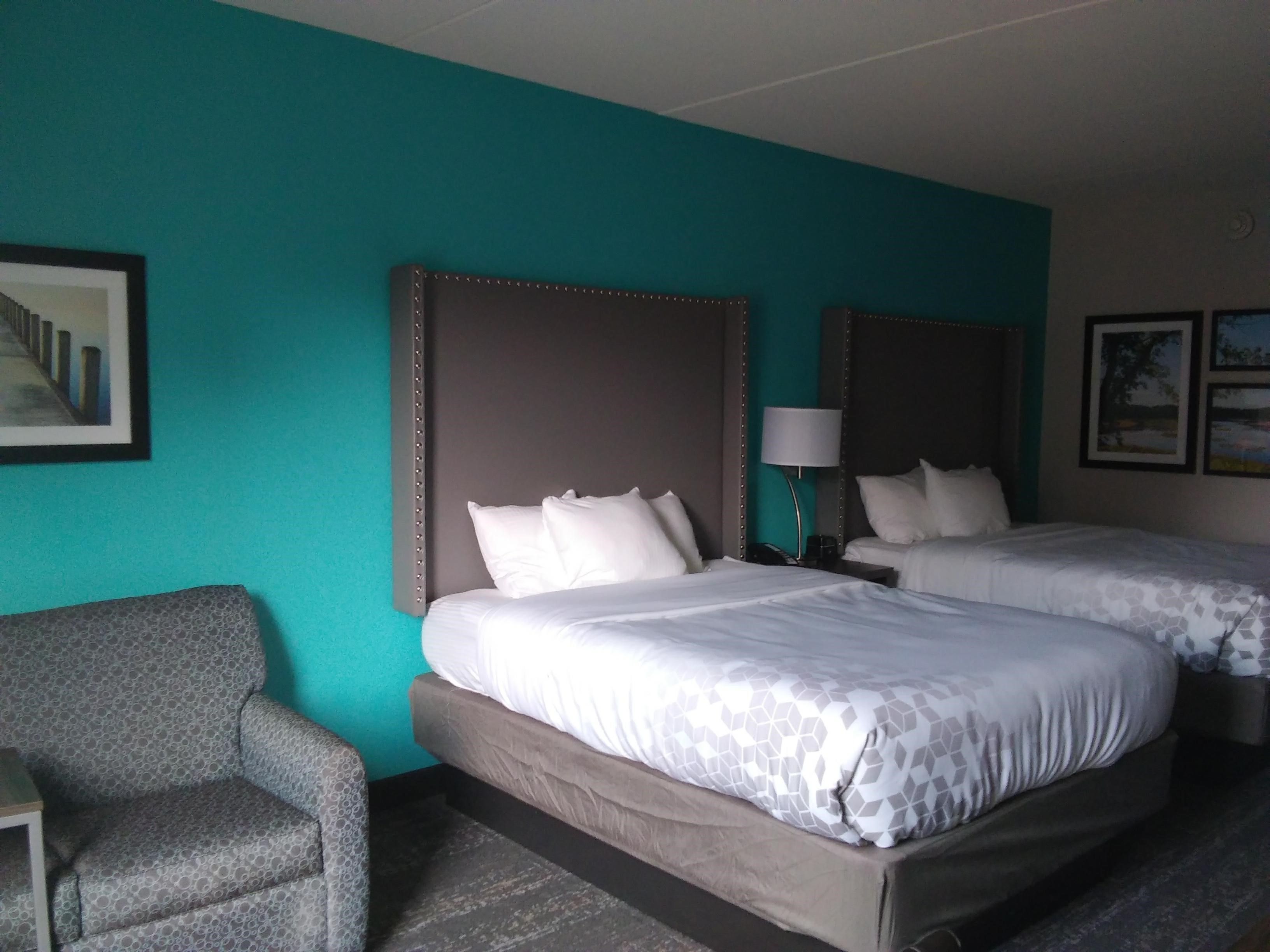 A hotel room with two beds, a grey armchair, and a small table.