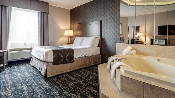 A hotel room with a bed, a large mirror, a bathtub, and a window with curtains. - Monte Carlo Inn - Brampton Suites (Brampton)