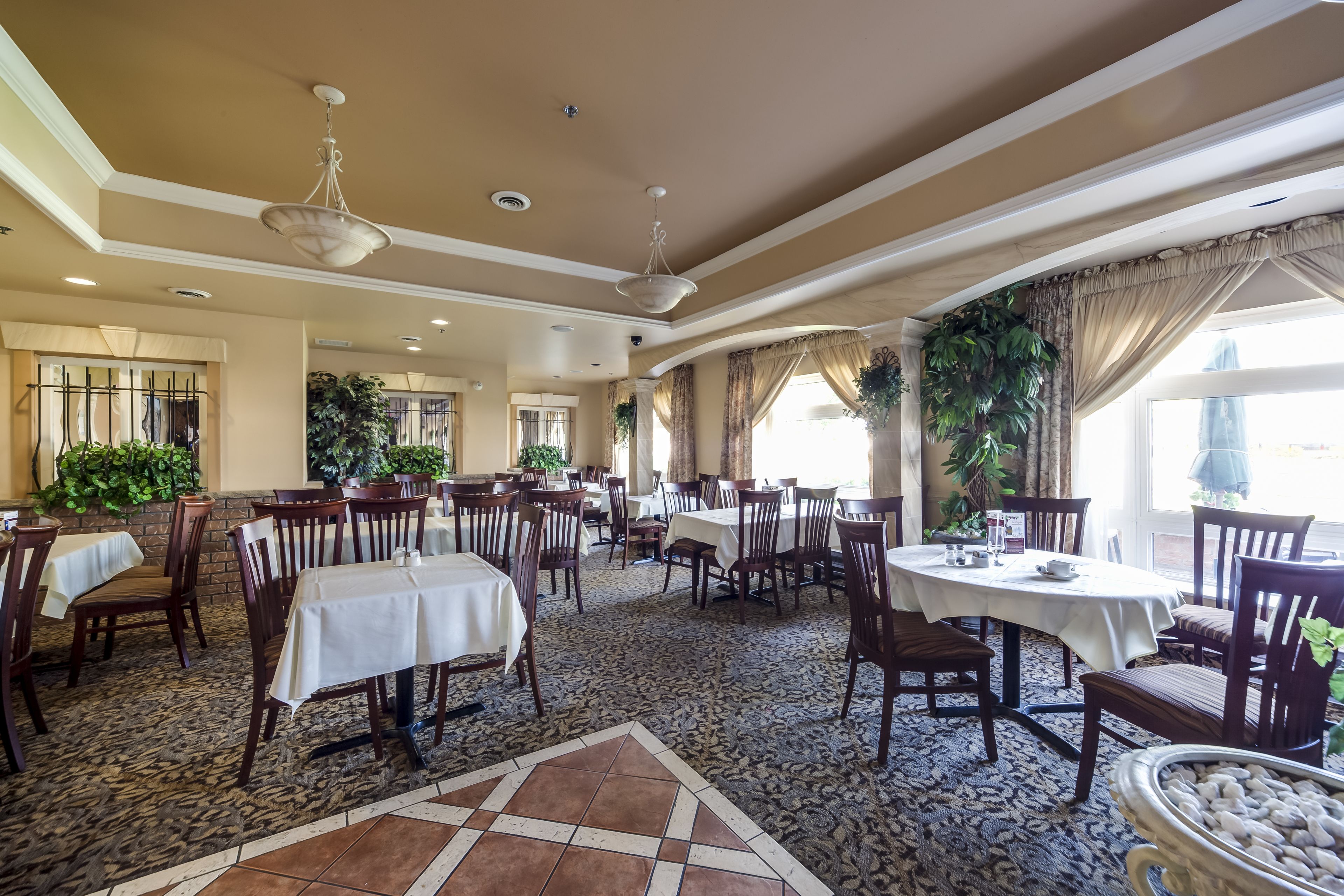 A spacious dining area with multiple tables and chairs, large windows with curtains, and decorative plants.