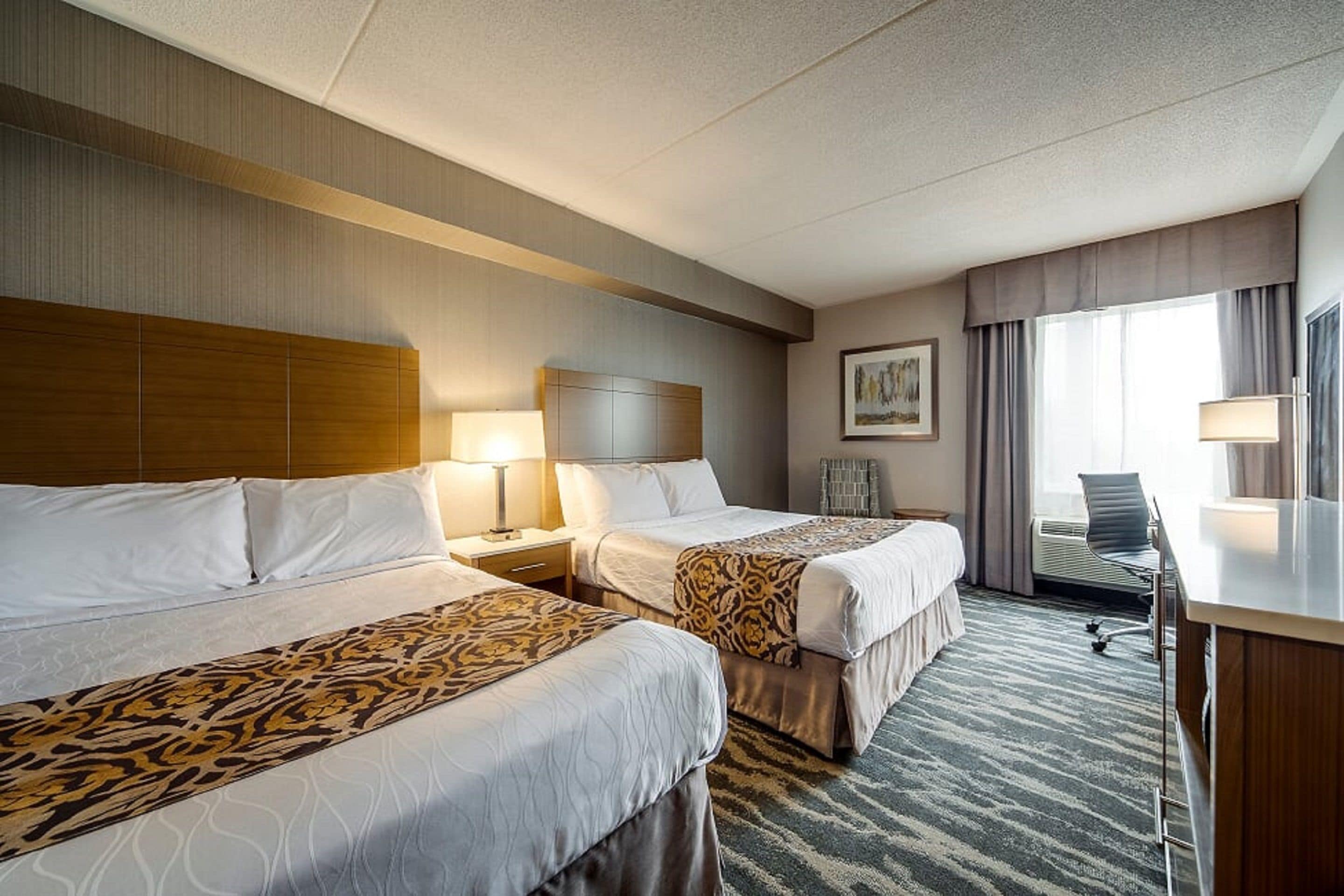 A hotel room with two beds, a desk, a chair, a lamp, and a window with curtains.