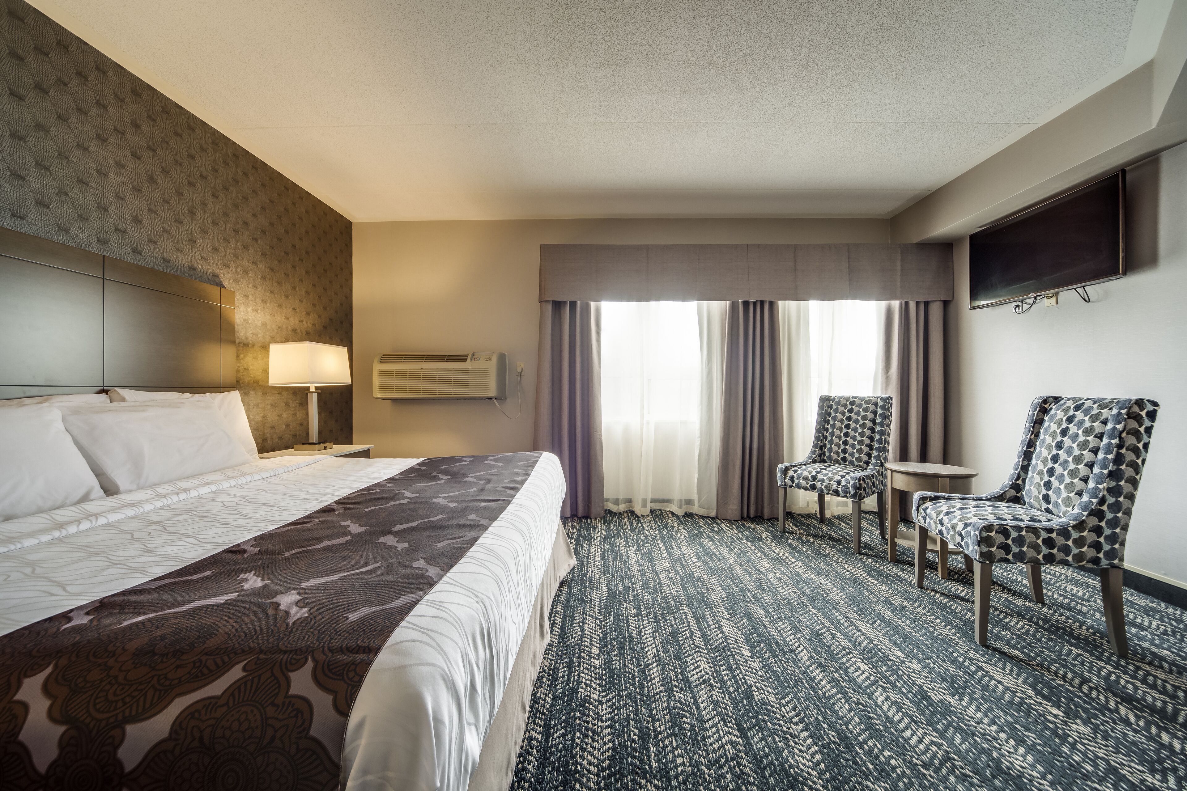 A hotel room with a large bed, two chairs, a TV, and a window with curtains.