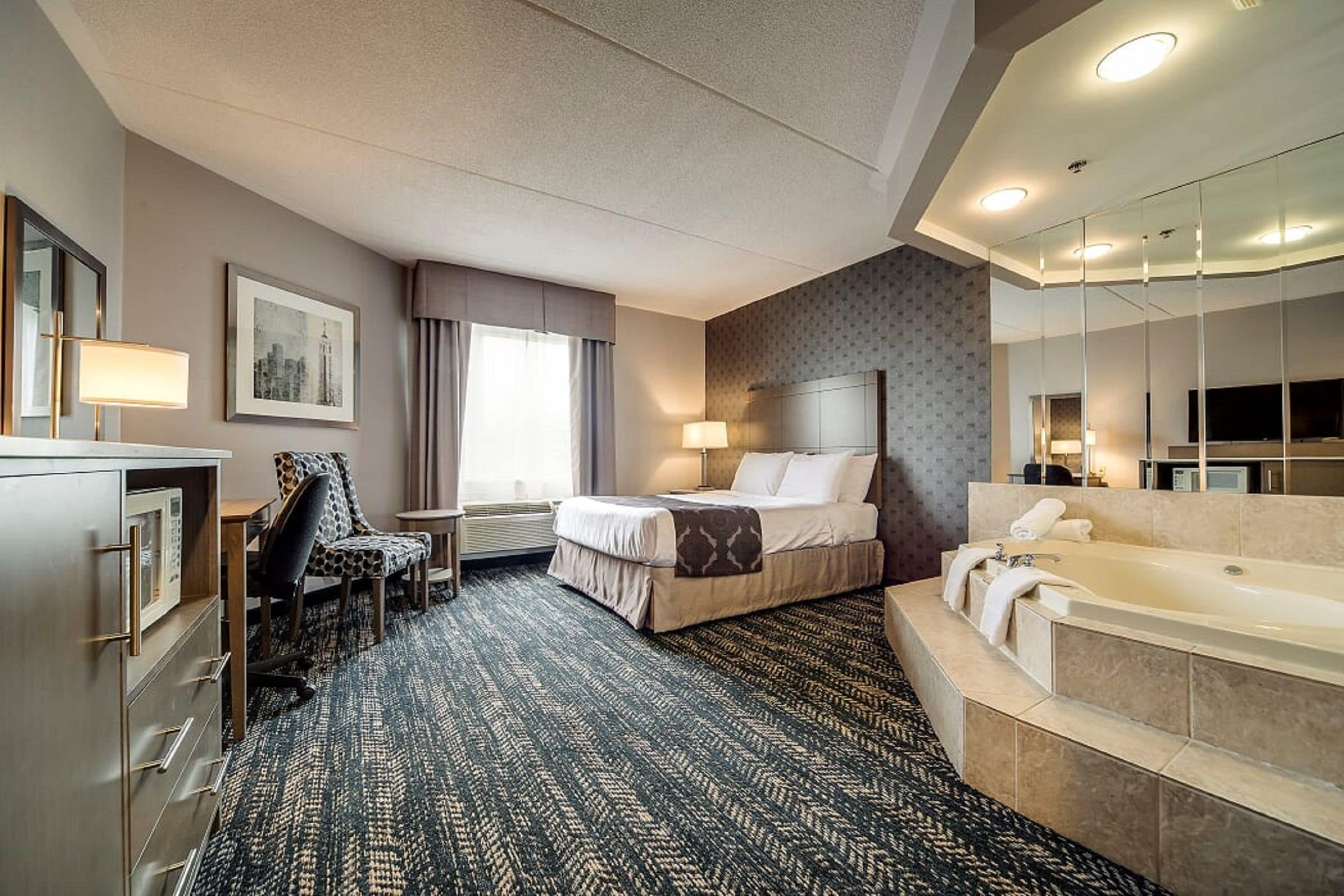 Una habitación de hotel con cama, escritorio con silla, baño con bañera y un espejo grande.