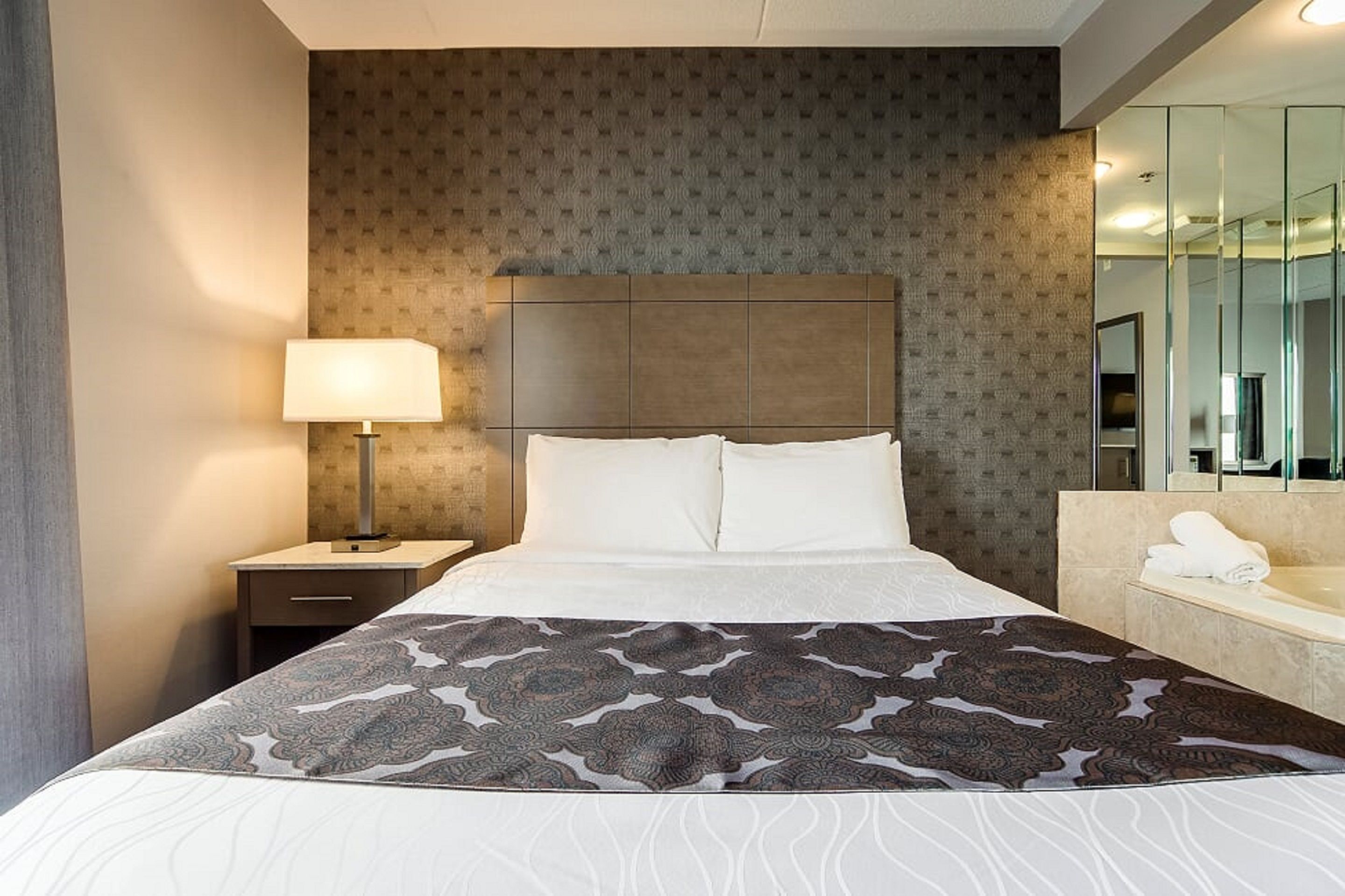 Habitación de hotel con una cama grande, una mesita de noche con lámpara, baño con espejo y colcha estampada.