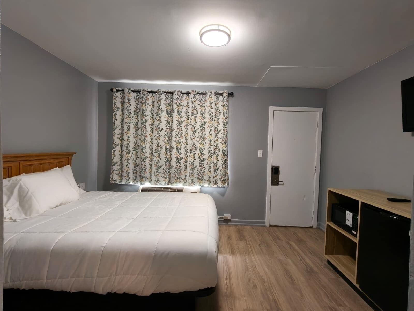 Une chambre à coucher avec un lit, une fenêtre avec des rideaux, une porte et un téléviseur fixé au mur.