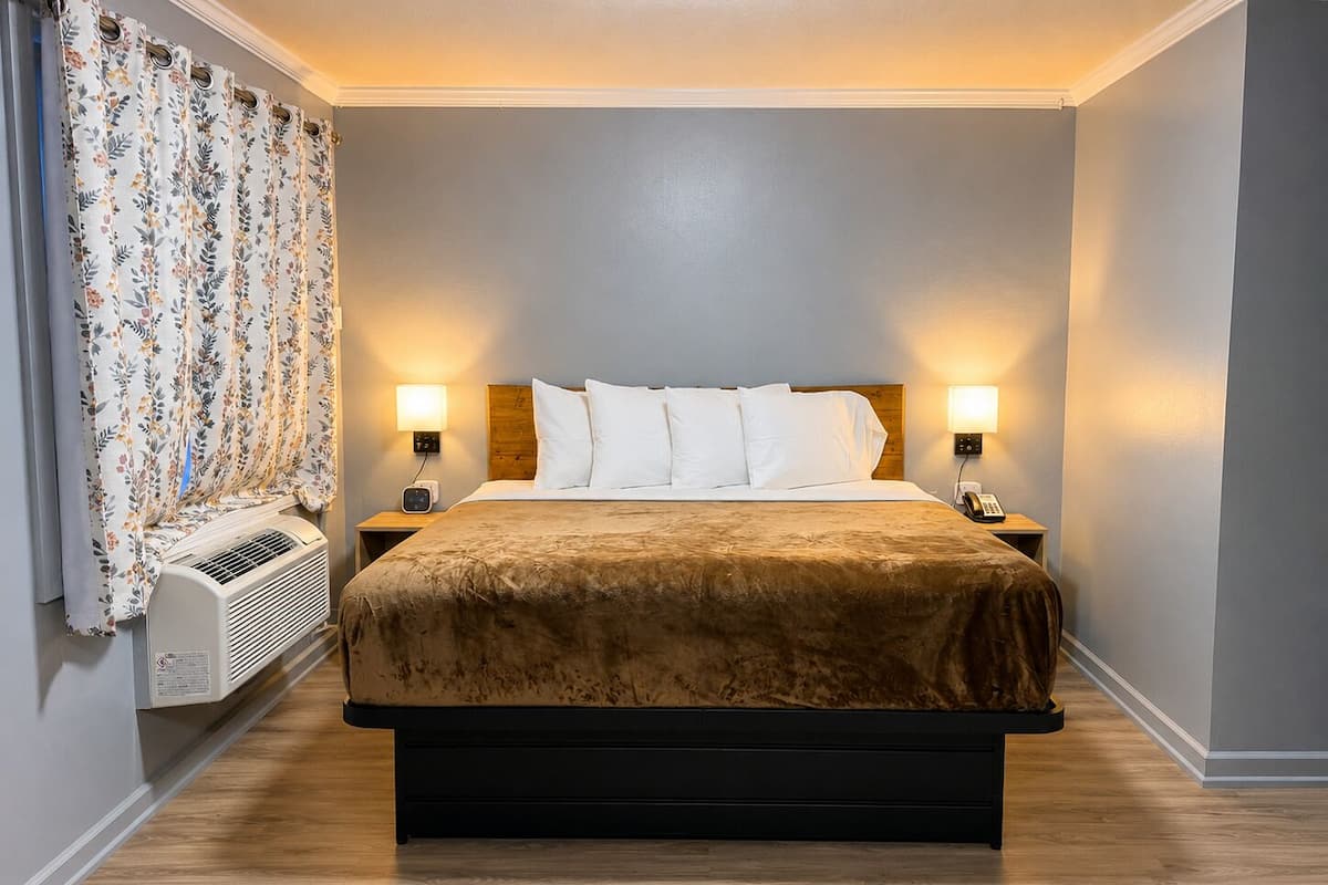 A hotel room with a bed, two bedside lamps, a heater, and a window with curtains.