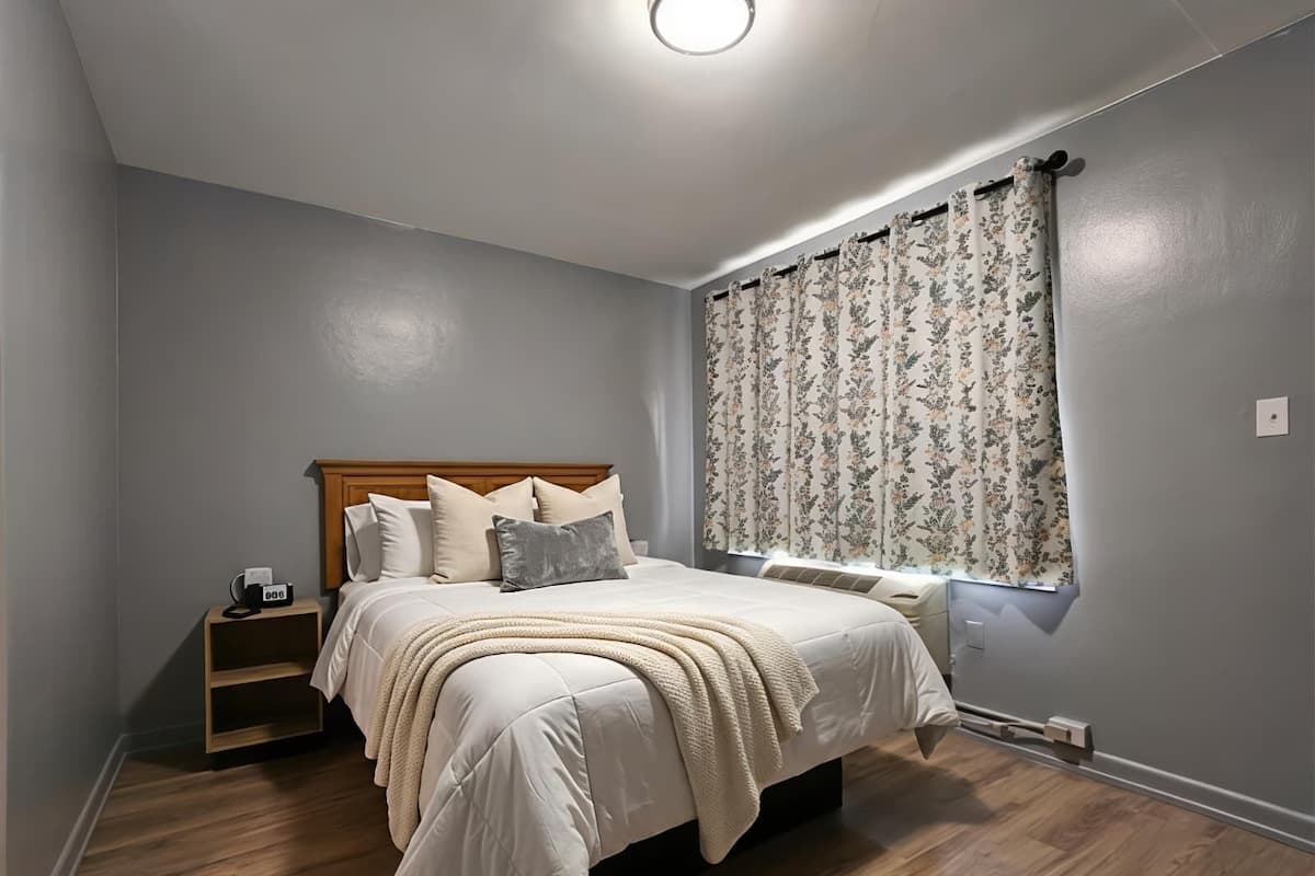 A bedroom with a bed, a nightstand, a window with floral curtains, and a wooden floor.
