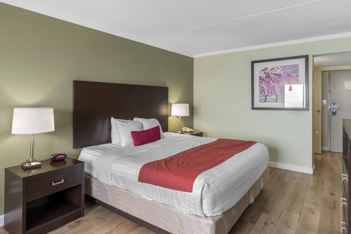 Habitación de hotel con una cama grande, dos mesitas de noche con lámparas, un tocador y un cuadro en la pared, además de una puerta al baño.