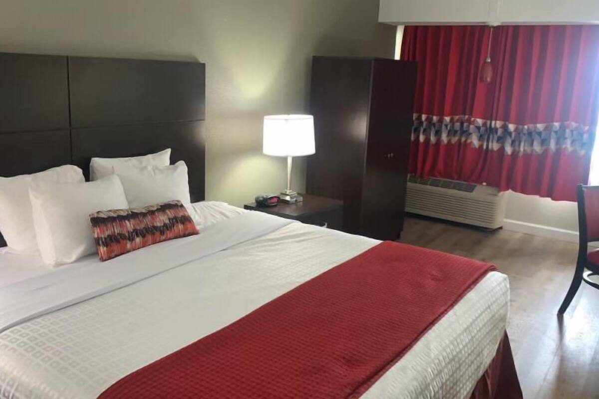 Una habitación de hotel con una cama grande, una mesita de noche con lámpara, una silla roja y una ventana con cortinas rojas.