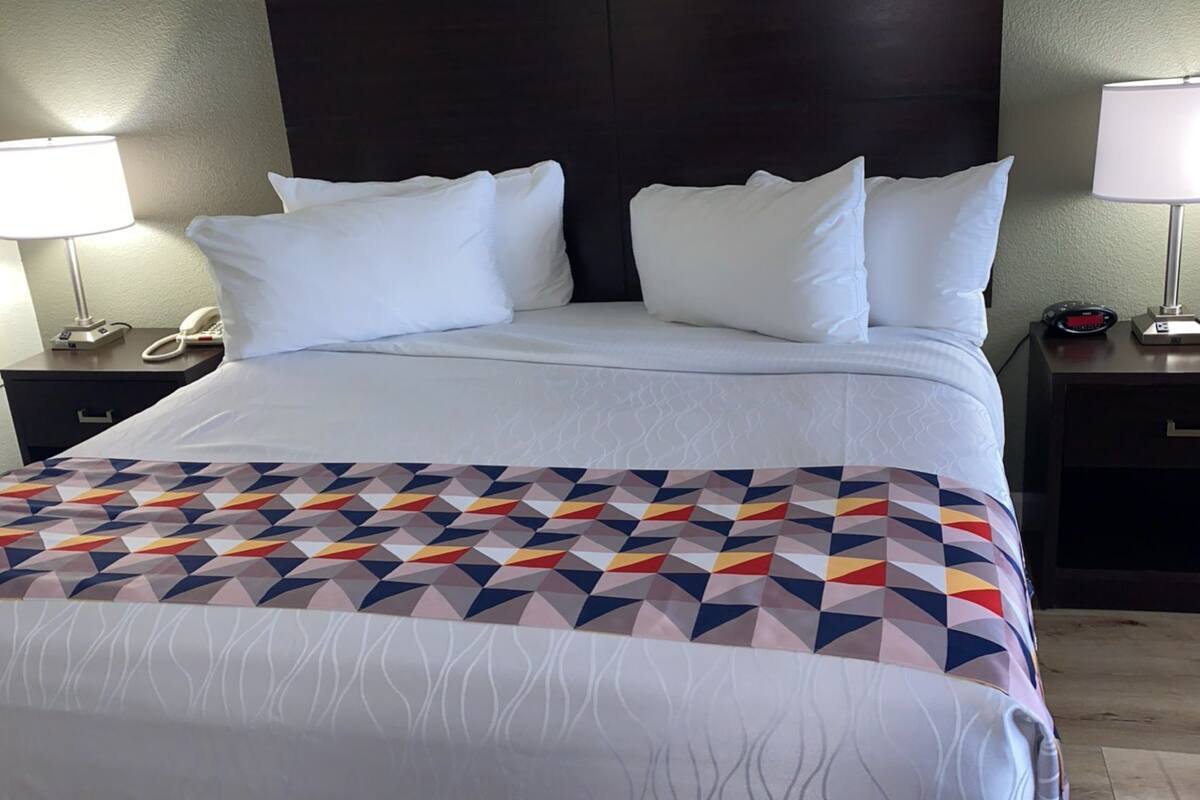 Una cama bien tendida con una manta de estampado geométrico, dos mesitas de noche con lámparas, un teléfono y un reloj pequeño en una de las mesitas.