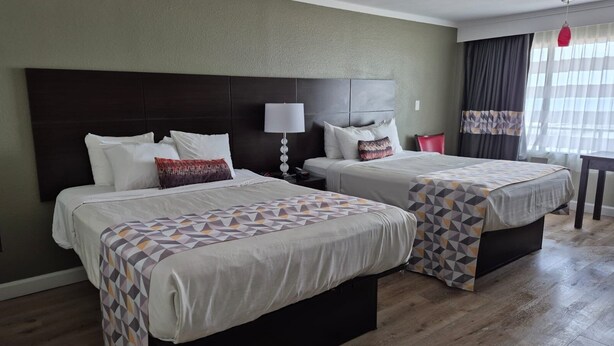 A hotel room with two beds, a dark headboard, a bedside table with a lamp, and a window with curtains. - Sandcastle Resort On The Beach (Virginia Beach)