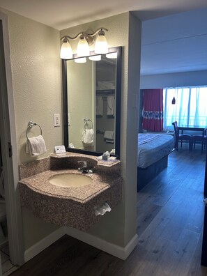 A bathroom with a granite sink, a mirror, and a vanity. There is a bed with a red curtain in the adjacent room. - Sandcastle Resort On The Beach (Virginia Beach)