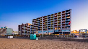A multi-story hotel with balconies, a sandy beach in front, and a pier shops sign in the background. - Sandcastle Resort On The Beach (Virginia Beach)