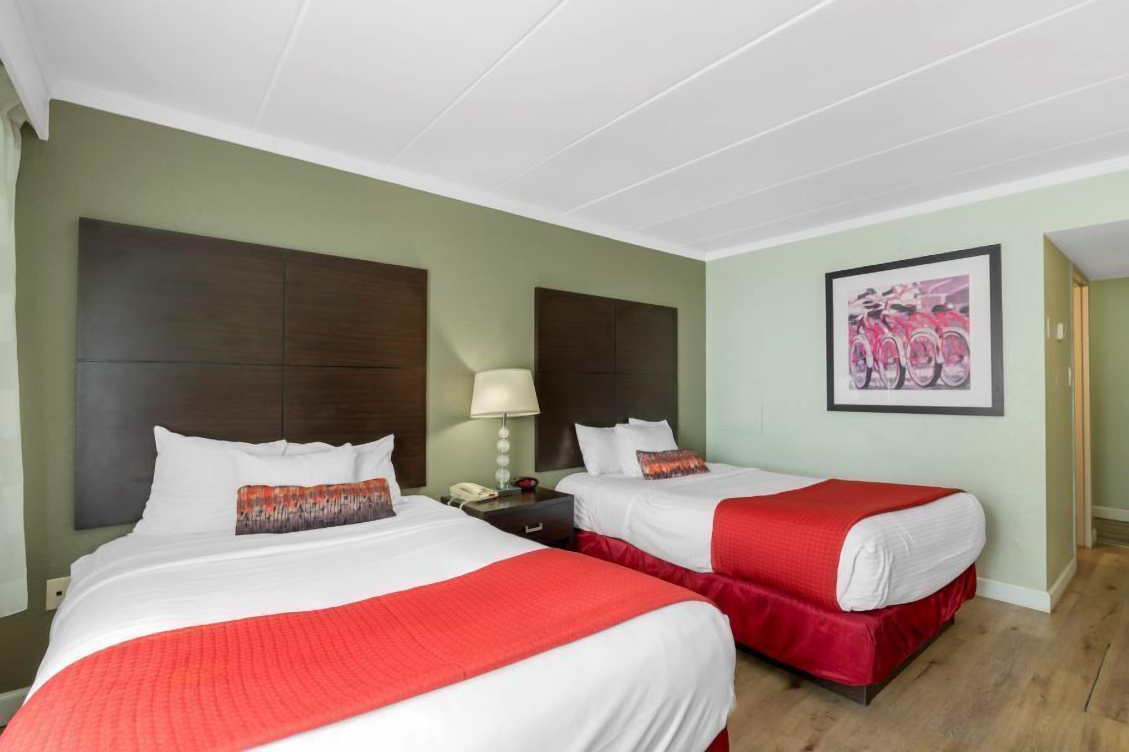 A hotel room with two beds, each with a red bedspread and white linens. There is a nightstand with a lamp and a phone, and a framed picture on the wall.