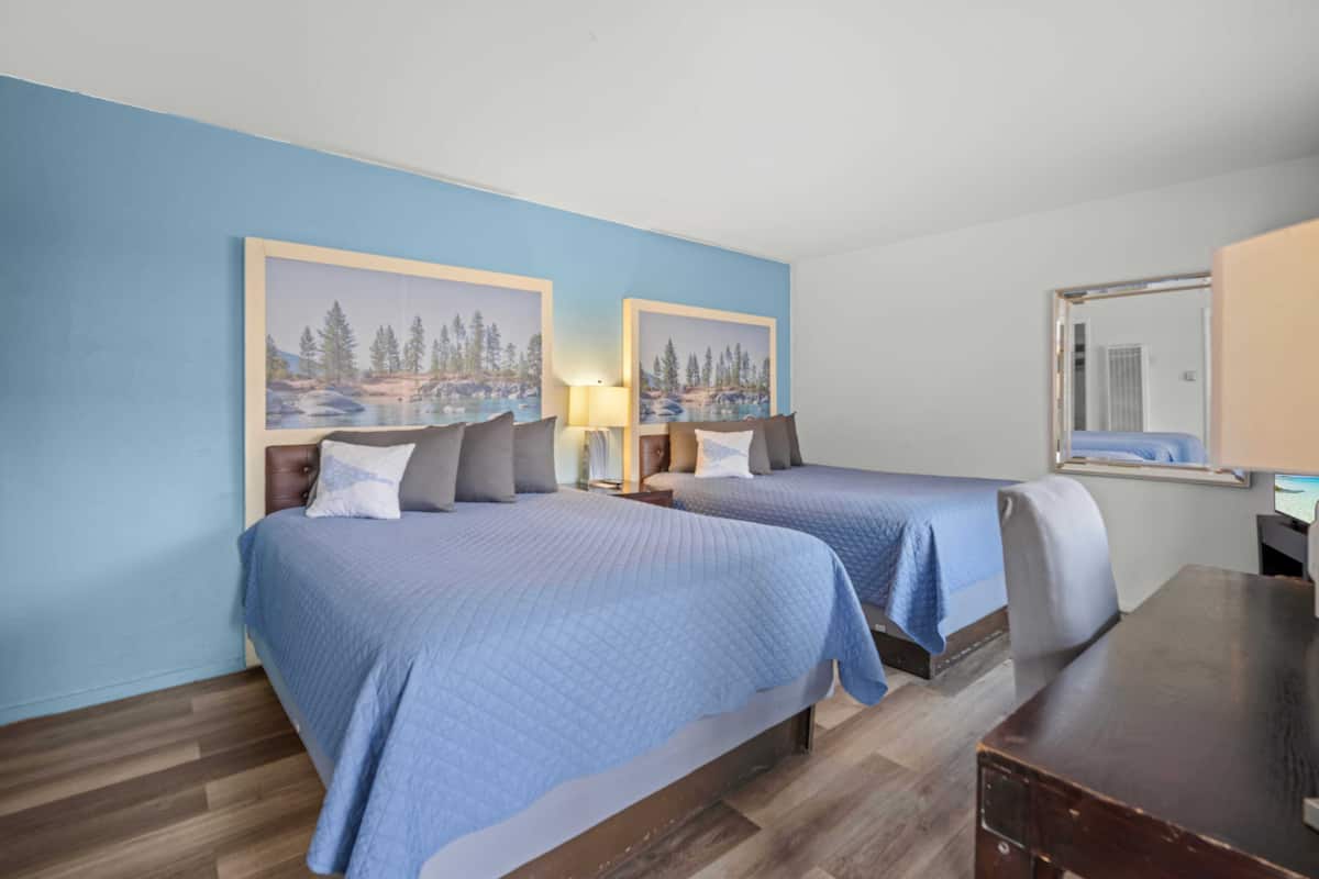 A hotel room with two beds, a wooden desk, a mirror, and a painting of a forest scene on the wall.