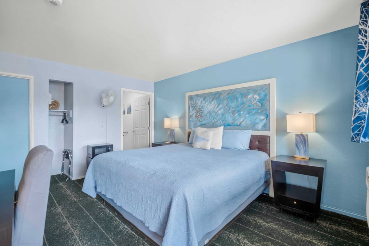 A hotel room with a large bed, two bedside tables with lamps, a wall-mounted artwork, a wardrobe, and a small seating area.
