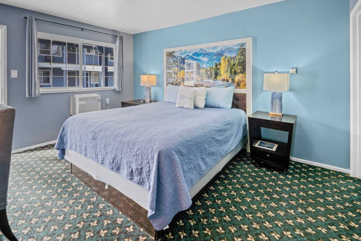 A bedroom with a large bed, two bedside tables with lamps, a window with curtains, and a painting on the wall.