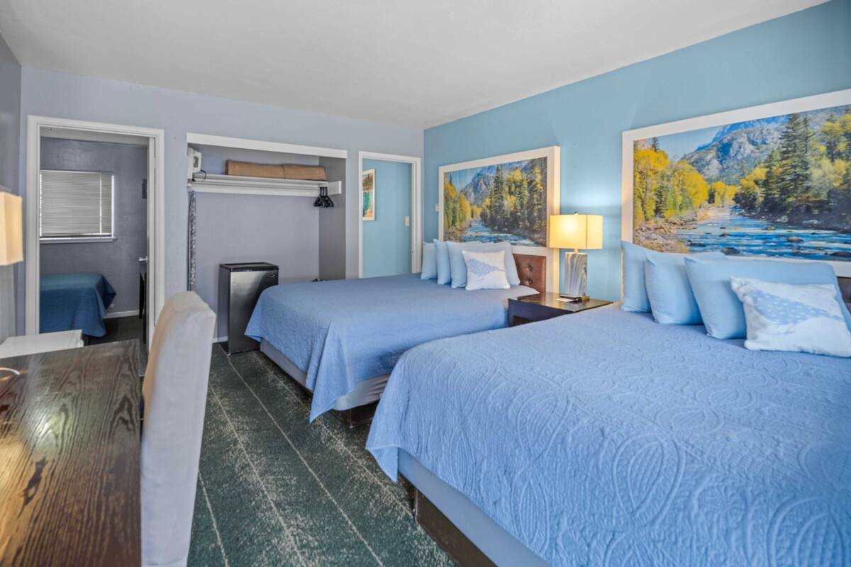 A hotel room with two beds, a desk, and a chair. There are paintings on the walls and a window with blinds.