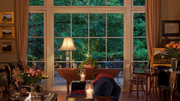 Ein gemütliches Zimmer mit einem großen Fenster, einem Klavier, einem Tisch mit Blumenvase und einem Sessel.