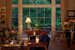 A cozy room with a large window, a piano, a table with a vase of flowers, and a chair. - The Stockade Bed and Breakfast (Baton Rouge)