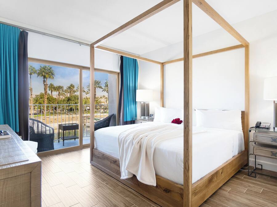 A four-poster bed with a view of palm trees and a pool, a desk with a remote control, and a nightstand with a lamp.