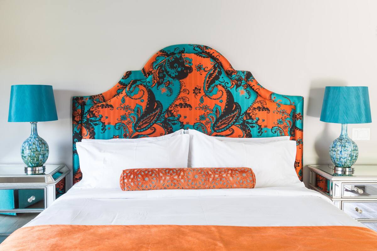 A bed with a patterned headboard, white bedding, and an orange throw blanket. There are two bedside tables with lamps and decorative vases.