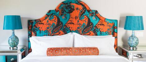 Una cama con cabecera estampada, ropa de cama blanca y una manta naranja. Hay dos mesitas de noche con lámparas y jarrones decorativos.