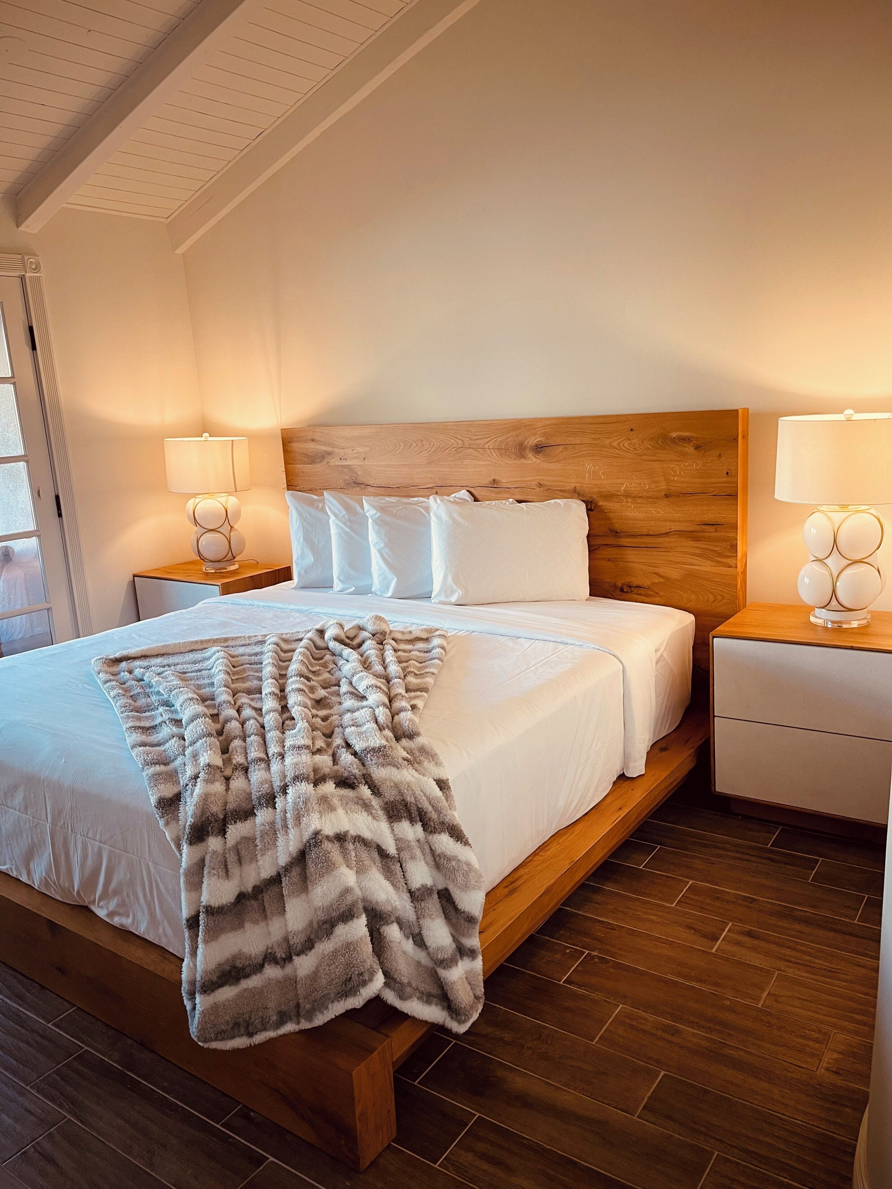 A modern bedroom with a wooden headboard, a large bed with white linens, two bedside tables with lamps, and a fluffy throw blanket on the bed.