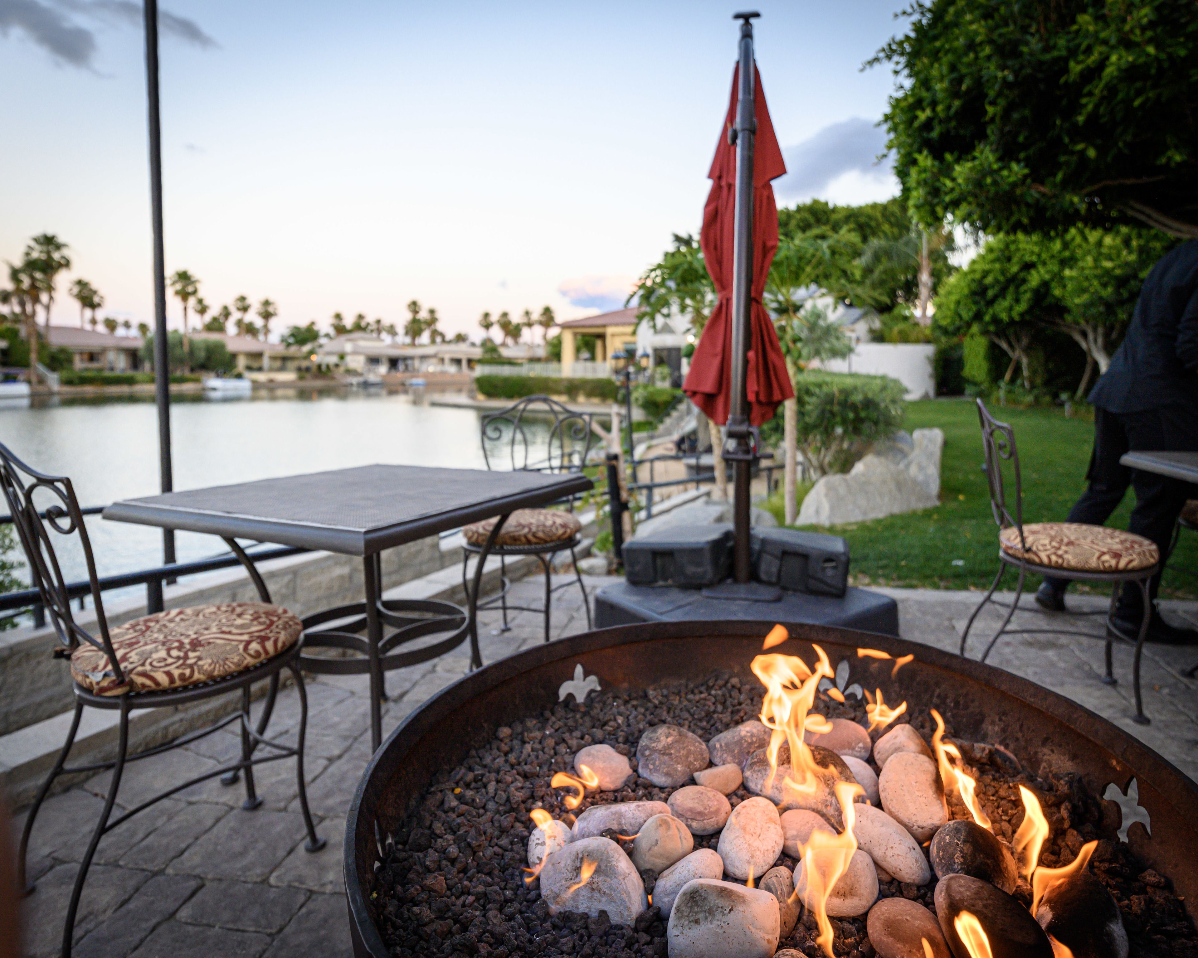 An outdoor seating area with a fire pit, tables, chairs, and a view of a body of water.