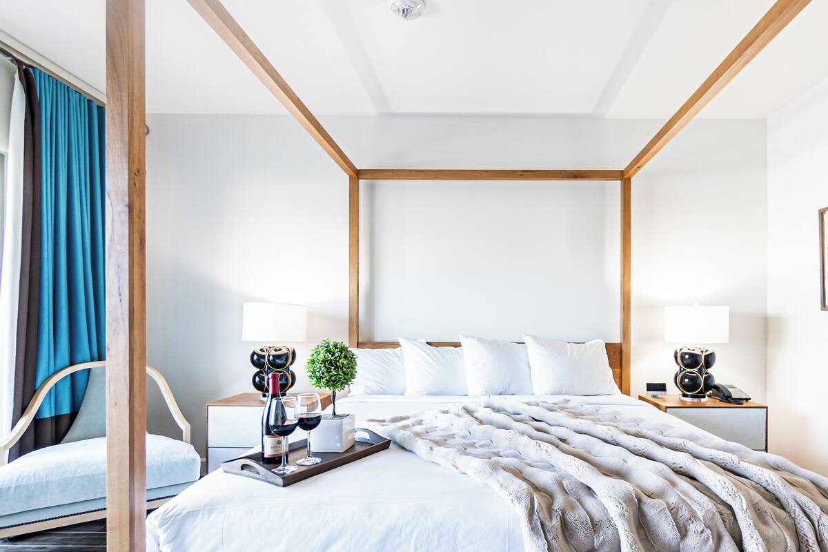 A modern hotel room with a four-poster bed, two bedside tables with lamps, a small potted plant, and a tray with wine glasses.