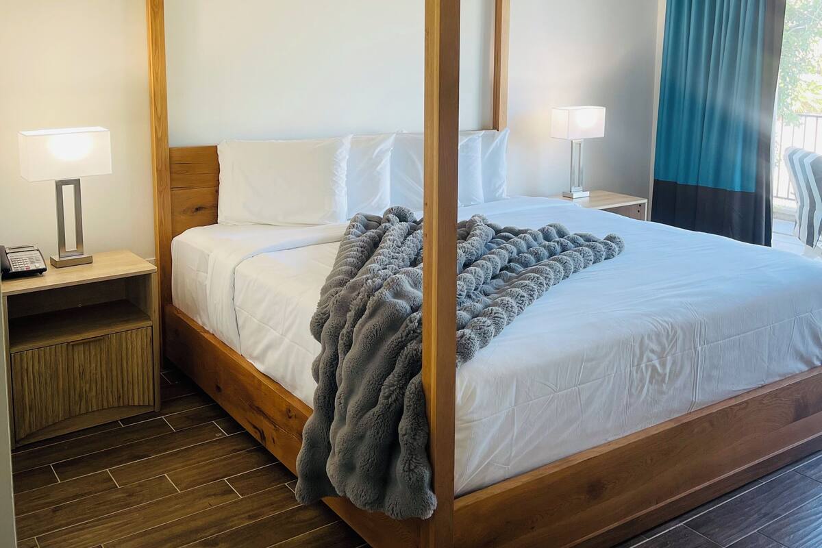 A four-poster bed with white linens, a grey throw blanket, and a wooden nightstand with a lamp.