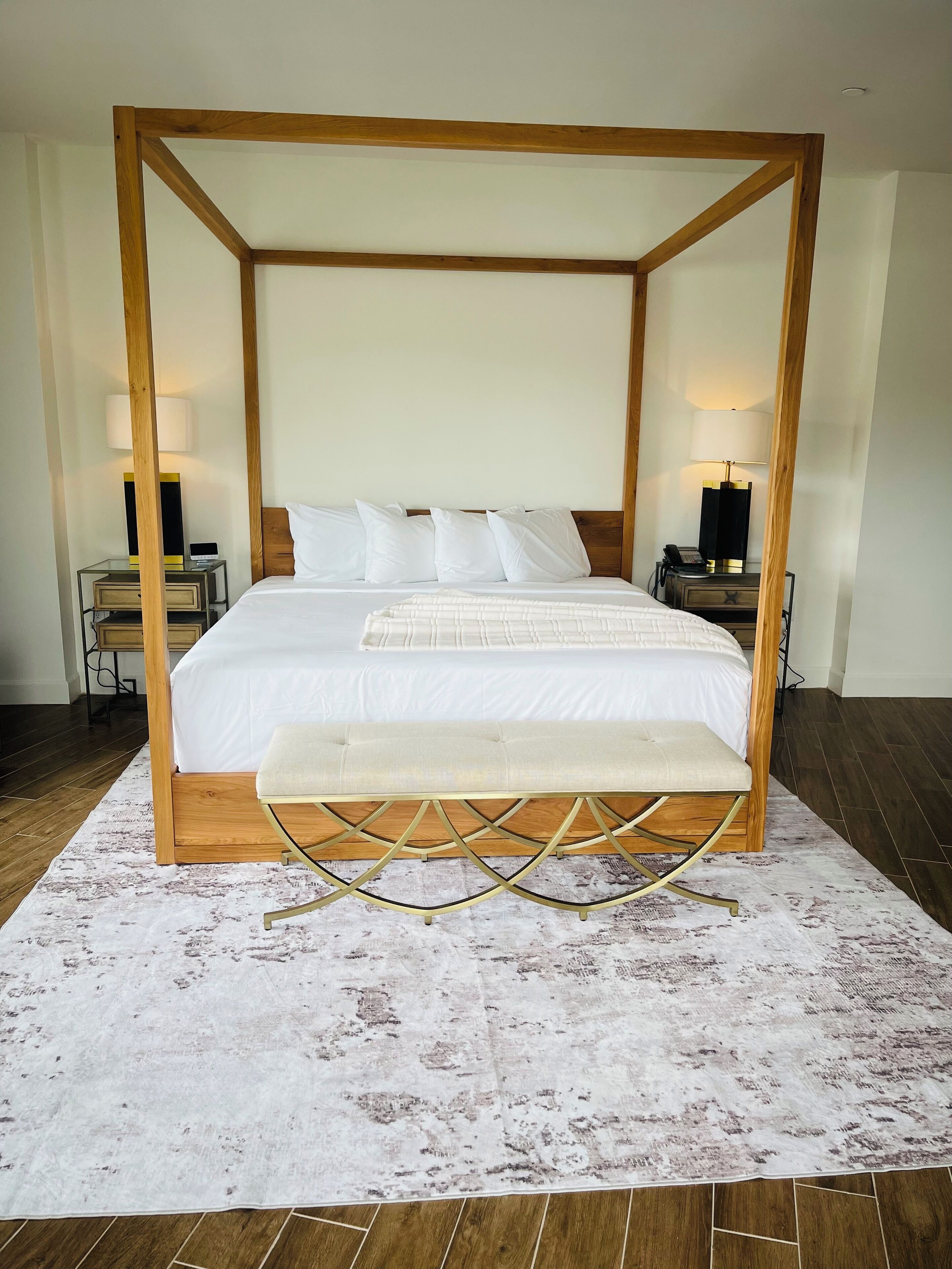A four-poster bed with a wooden frame, white bedding, and a bench at the foot. There are two bedside tables with lamps, and a patterned rug on the floor.