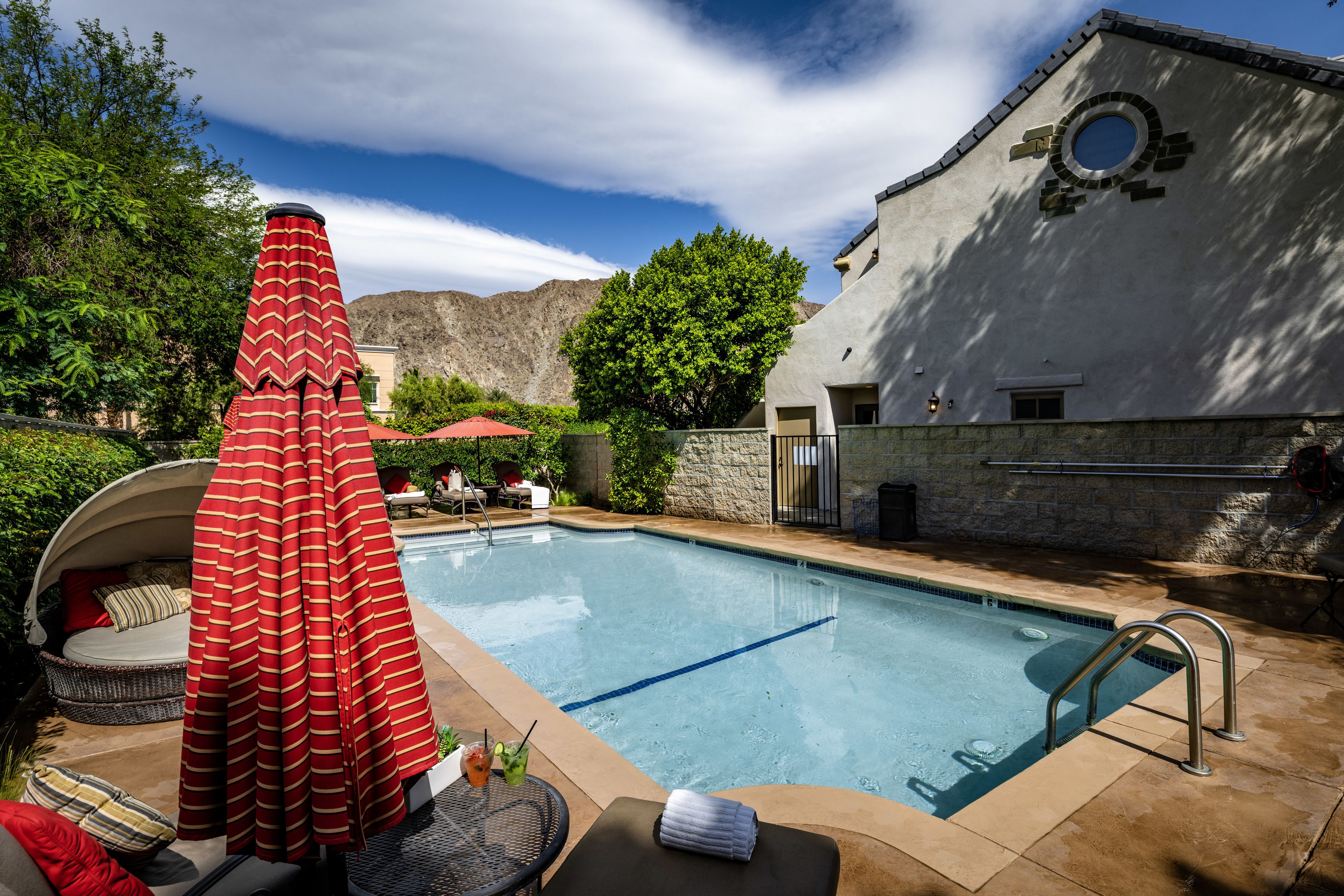 Une piscine avec un parasol rouge, des chaises longues et un mur en pierre.