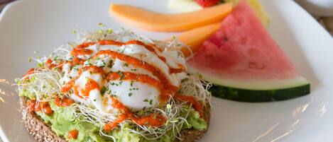 A plate with a sandwich topped with avocado, sprouts, and a poached egg, alongside slices of watermelon and a strawberry.