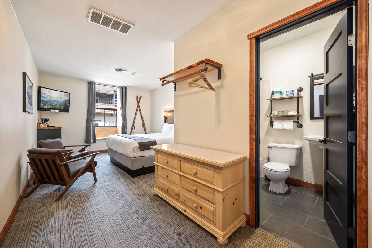 Quarto de hotel com uma cama, cômoda de madeira, cadeira, televisão e banheiro com vaso sanitário e prateleiras.