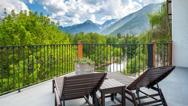 A balcony with a table and two chairs, overlooking a river and mountains. - Hotel Leavenworth (Leavenworth)