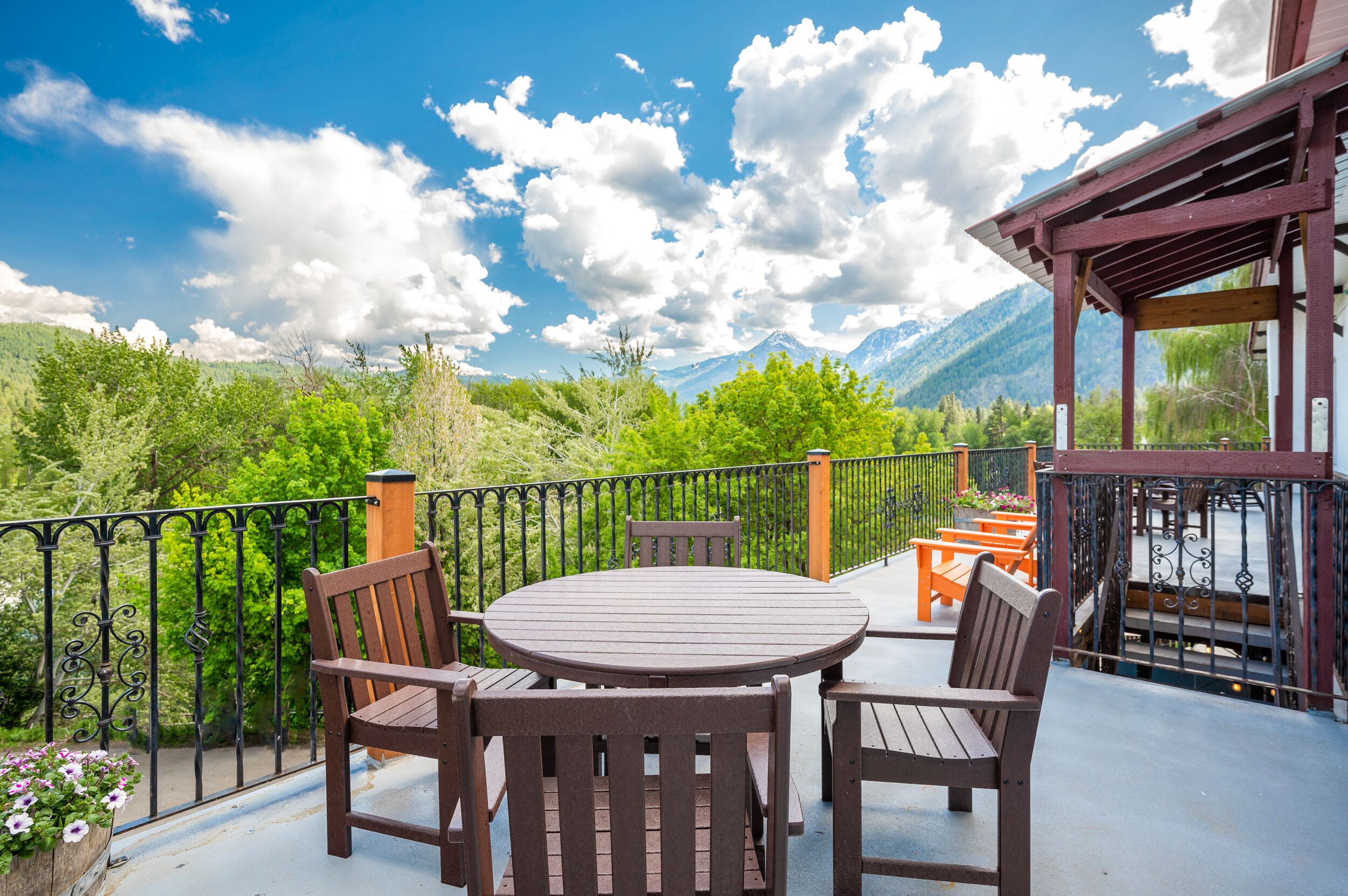 A terrace with a round wooden table and chairs, surrounded by a black wrought iron railing, overlooking a mountainous landscape.