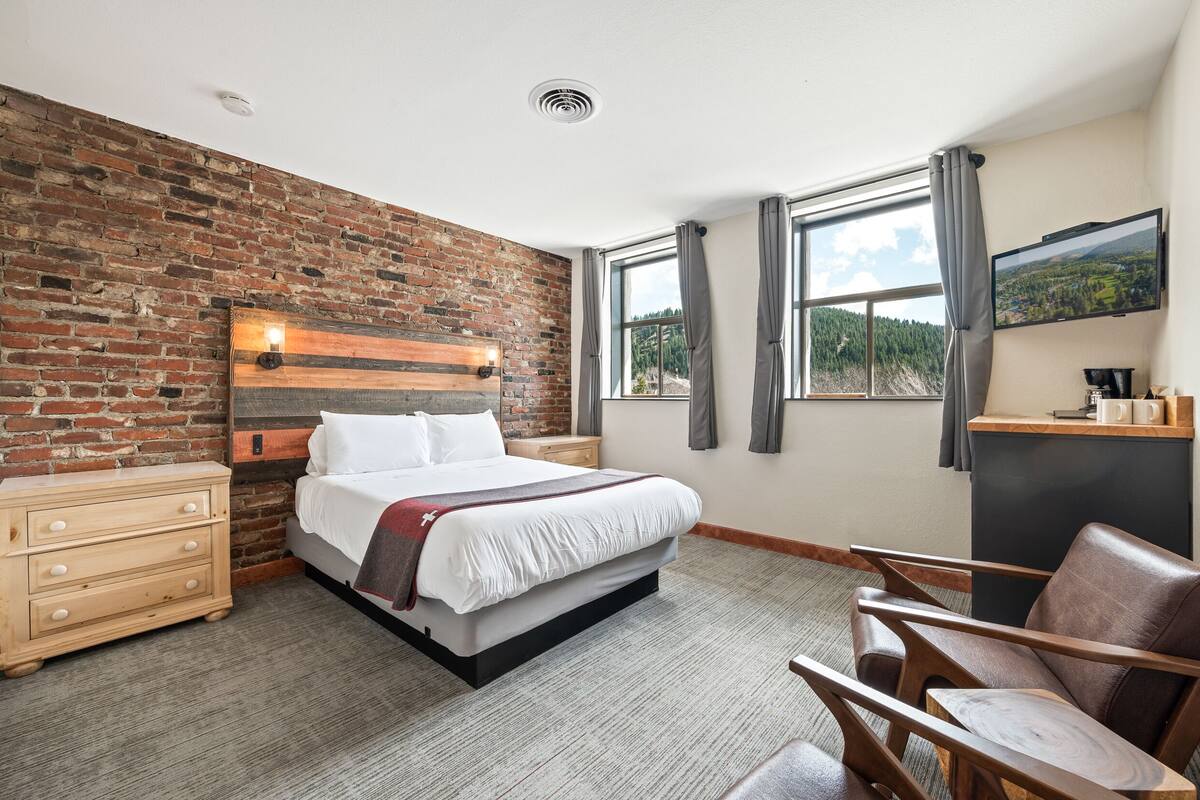 Quarto de hotel com parede de tijolos, cama com roupa de cama branca, penteadeira de madeira, escrivaninha com computador e vista para uma floresta pela janela.