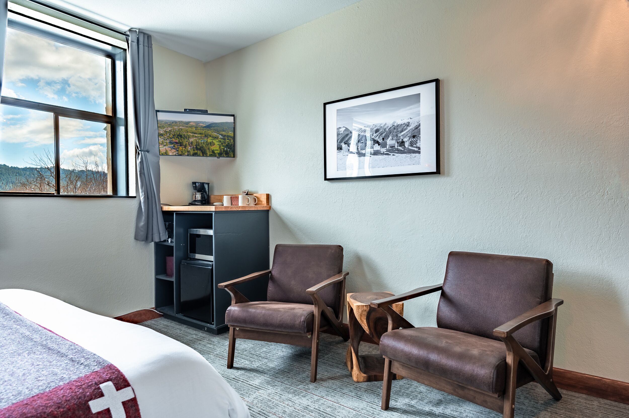A hotel room with a bed, two leather chairs, a small wooden table, a desk with a computer, and a framed picture on the wall.