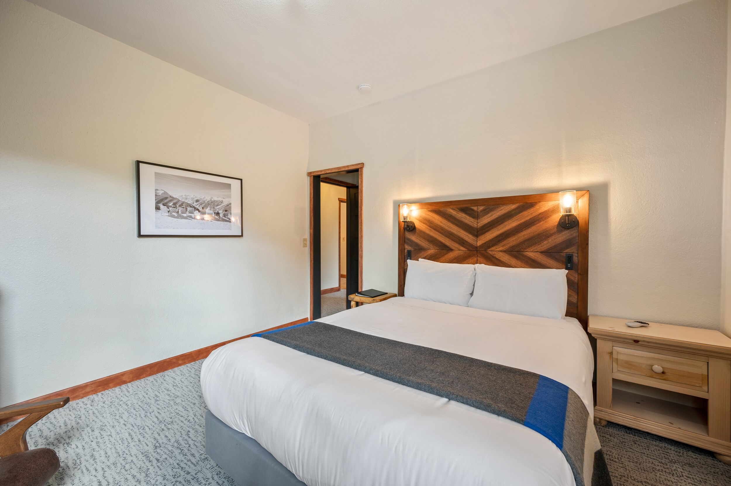 Quarto de hotel com uma cama grande, mesa de cabeceira, criado-mudo, quadro emoldurado na parede e cabeceira de madeira com luz.
