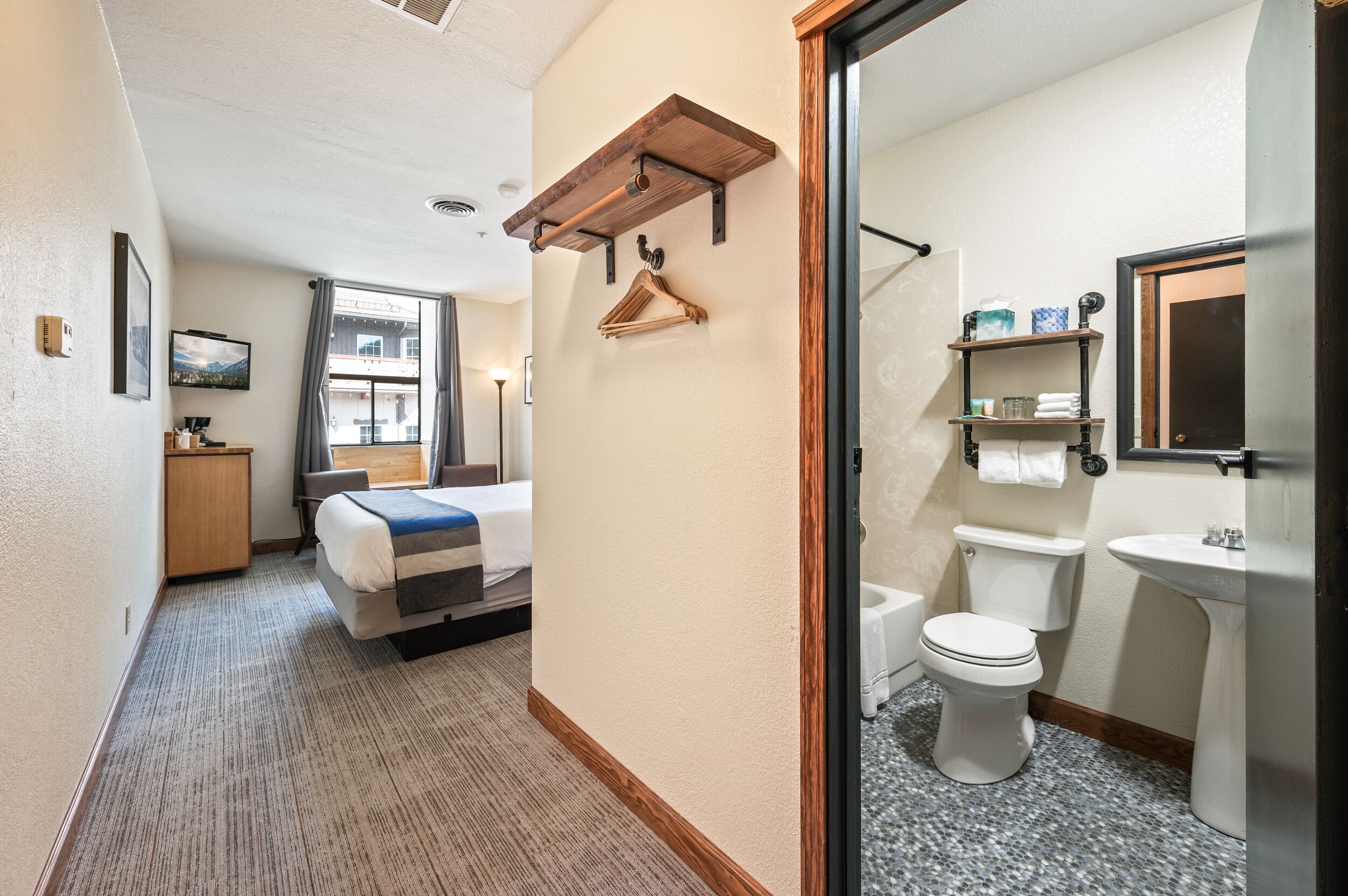 Camera d'albergo con un letto, una scrivania, una finestra, un bagno con il water e un lavandino, e una mensola in legno.