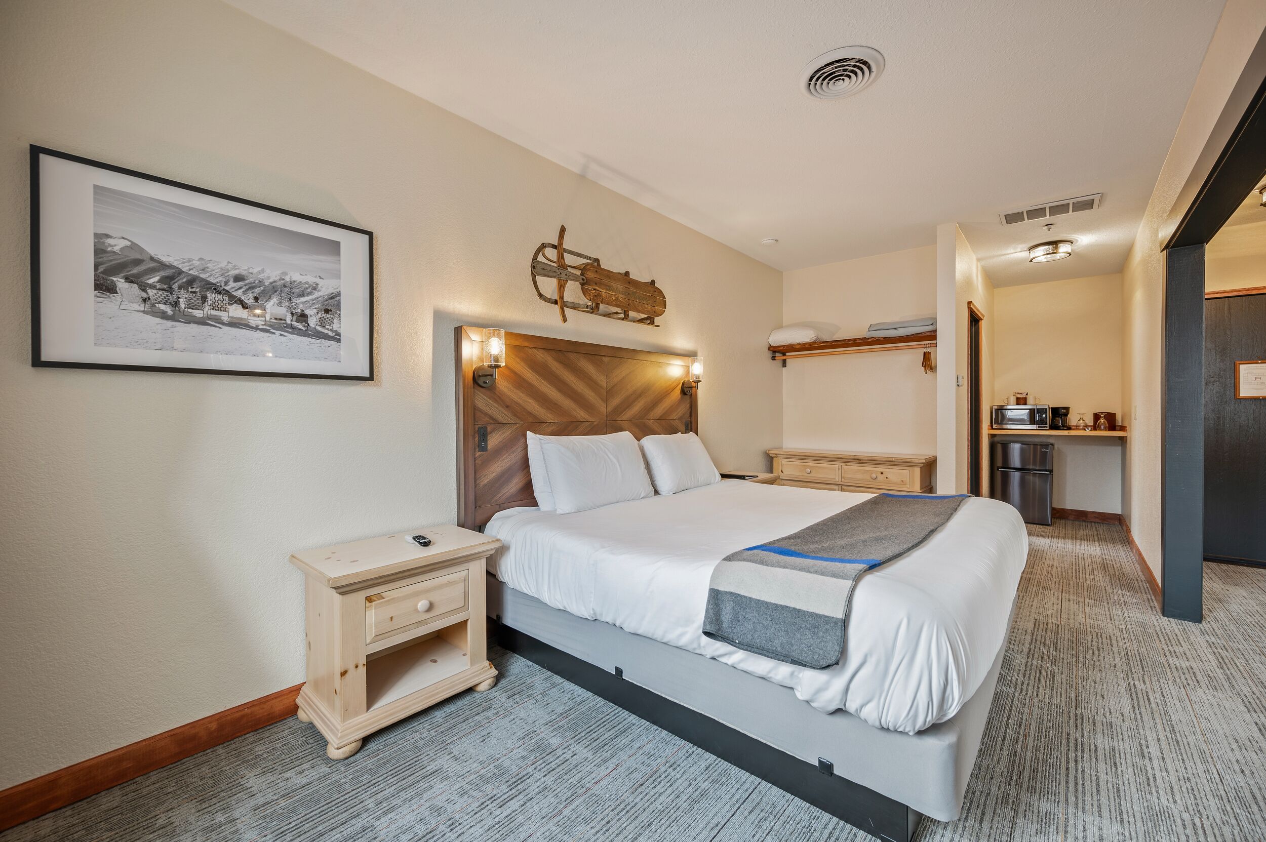 Ein Hotelzimmer mit Bett, Nachttischen, einem kleinen Schreibtisch, einem Kühlschrank und einem gerahmten Bild an der Wand.