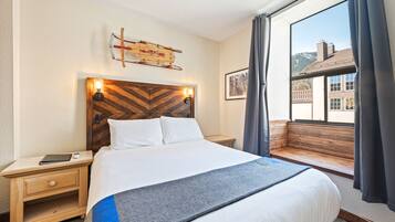 Quarto de hotel com uma cama grande, cabeceira de madeira, mesa de cabeceira com abajur, janela com vista para edifícios e uma obra de arte com tema de teleférico na parede.