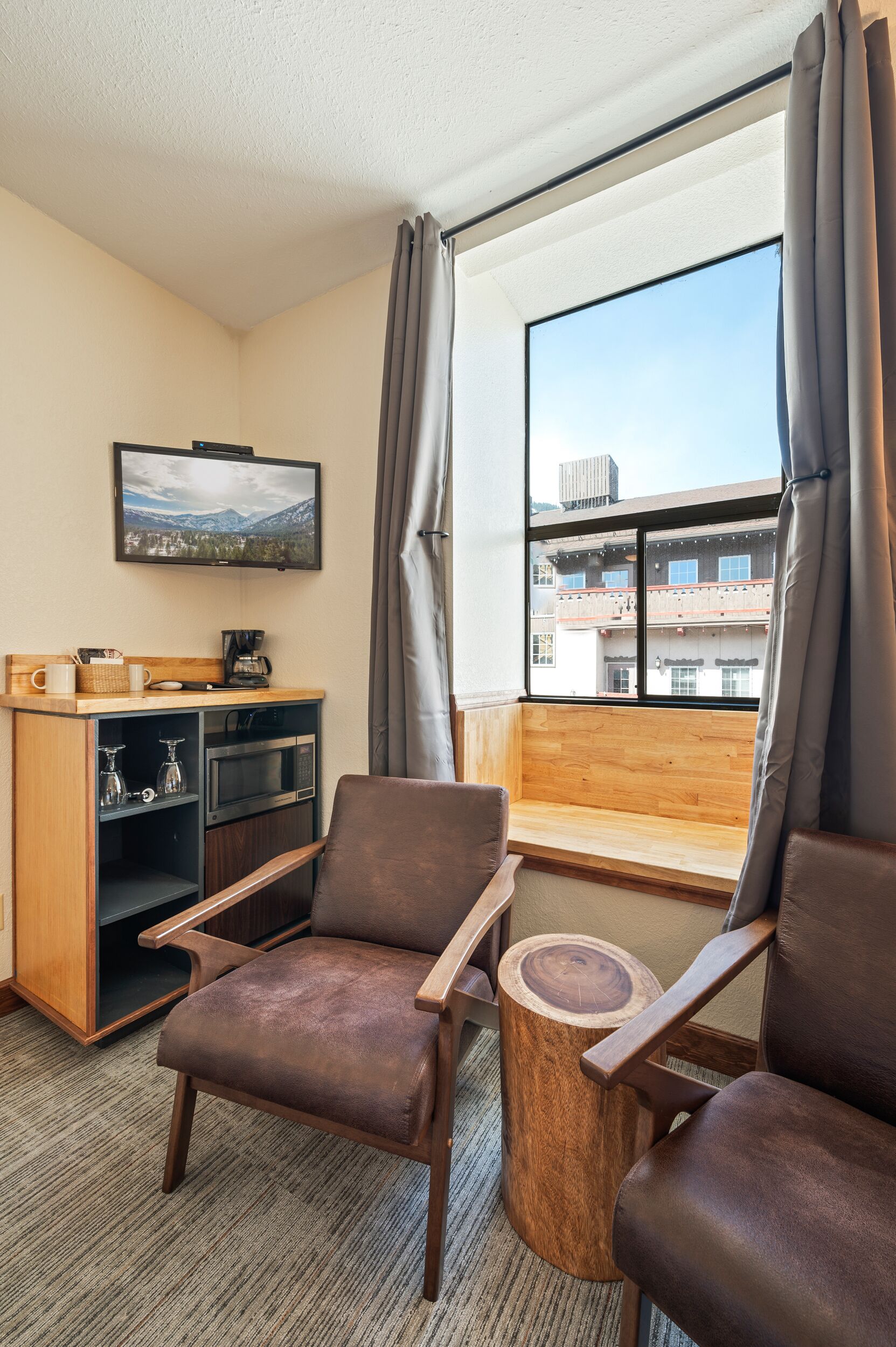 Habitación de hotel con un banco de madera, dos sillones de cuero marrón, una kitchenette pequeña, un televisor montado en la pared y un ventanal con vistas a edificios.