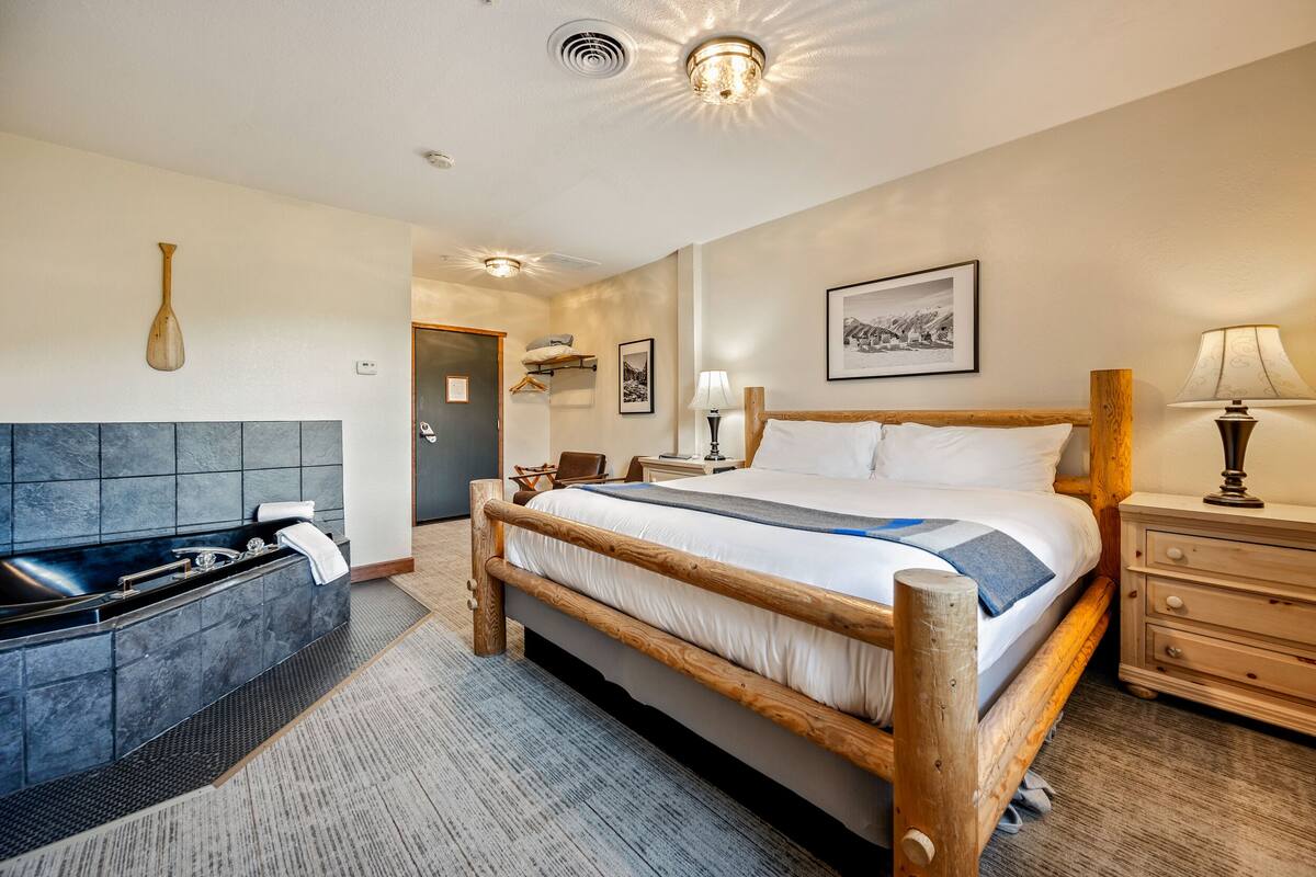 Quarto de hotel com cama de madeira, banheira, mesa de cabeceira com abajur e quadro pendurado na parede.