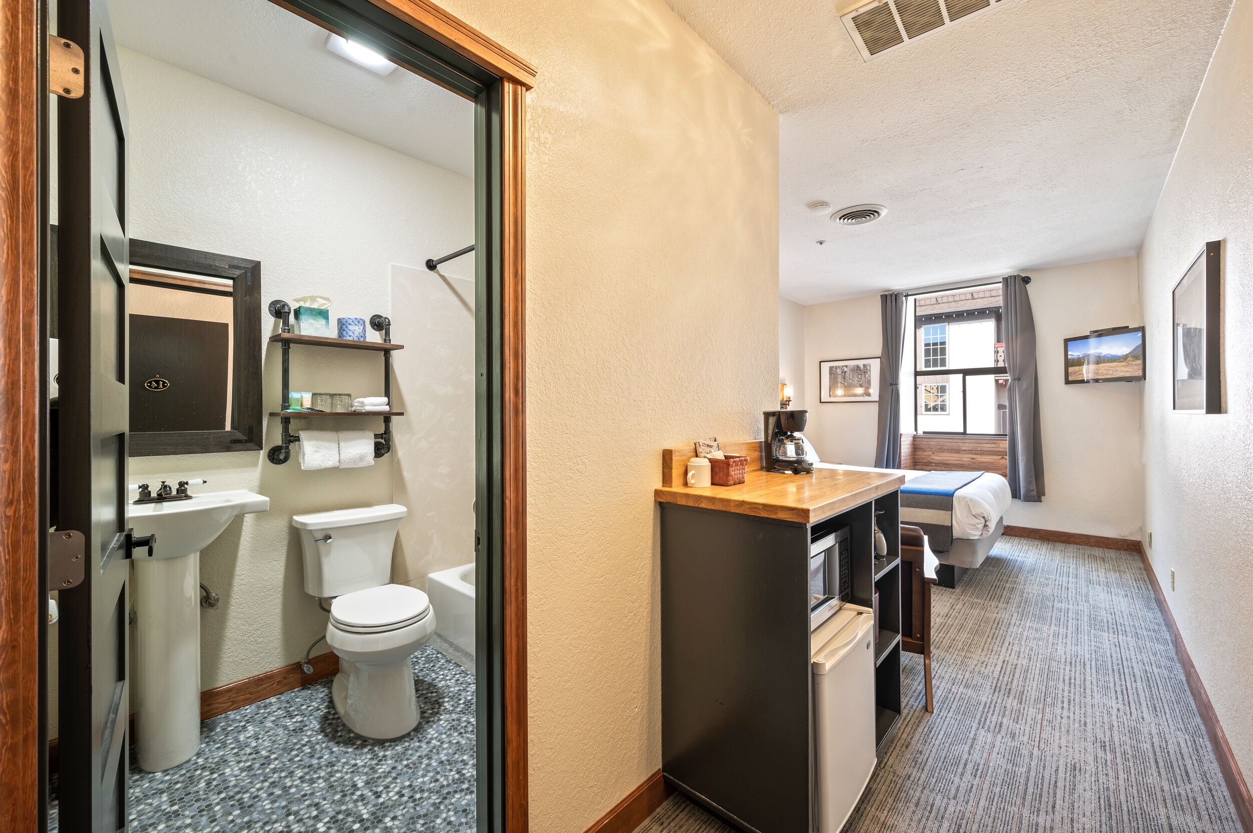 A compact hotel room with a bathroom featuring a sink, toilet, and mirror. A small kitchenette with a coffee maker and a bed with a window view is also visible.
