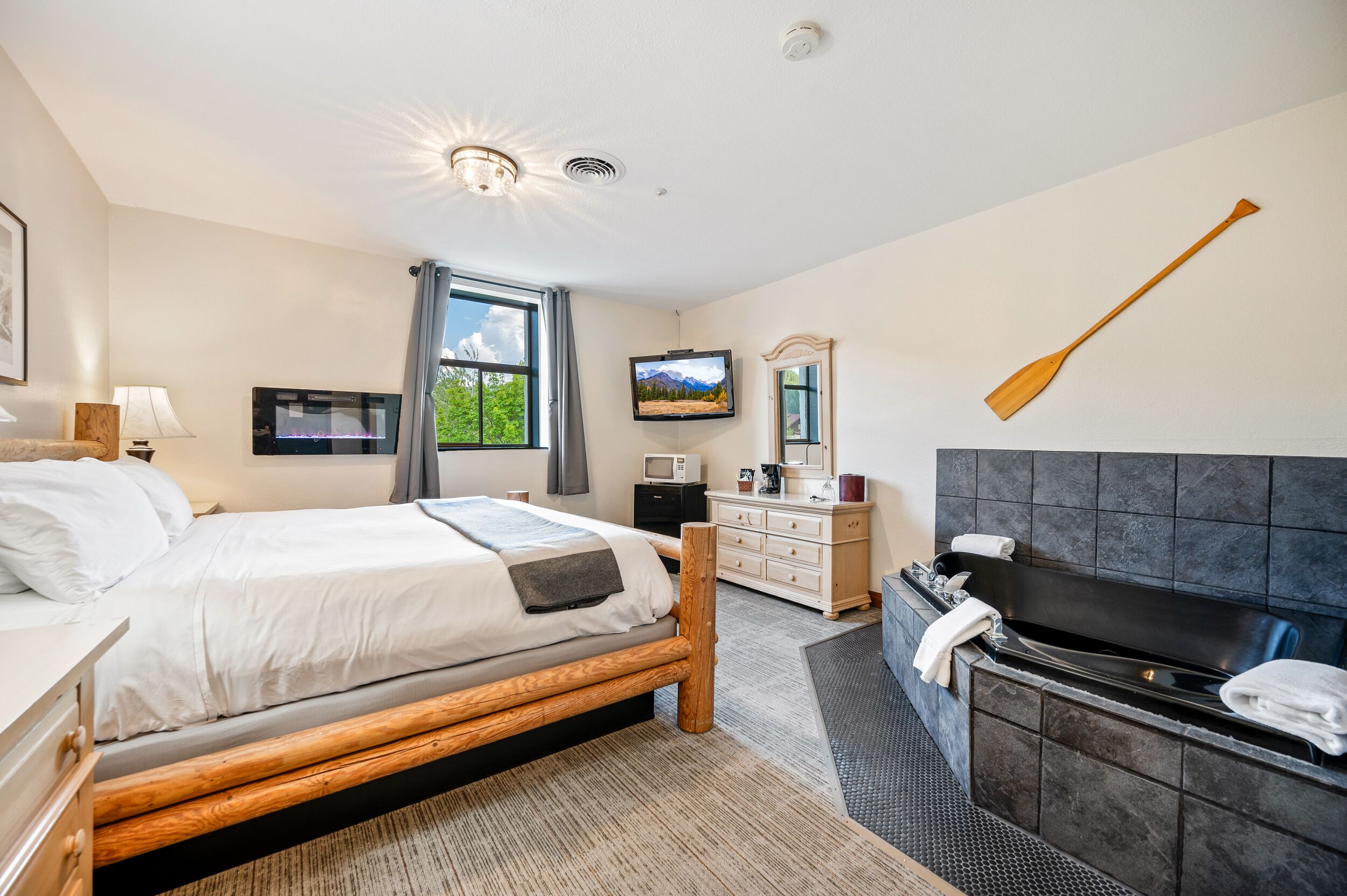 A hotel room with a bed, a TV, a desk, a dresser, a mirror, a bathtub, and a wooden paddle on the wall.